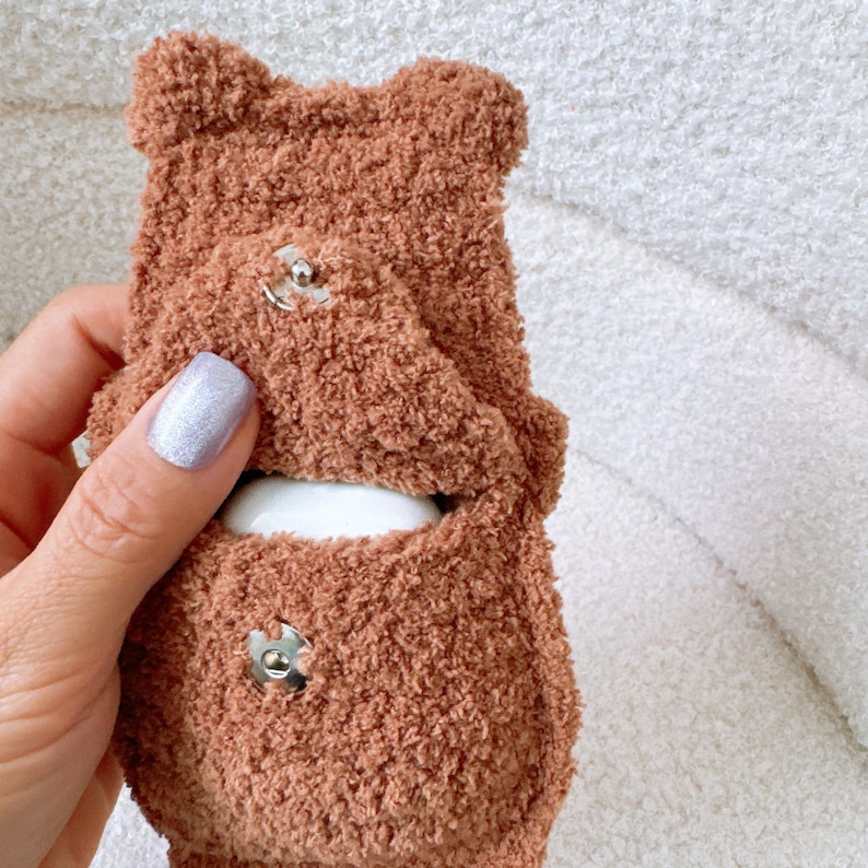 May include: A brown, fuzzy, teddy bear-shaped protective case for wireless earbuds. The case features a soft, textured exterior with two small rounded ears at the top and a front flap that secures with a silver-toned metal snap button closure.