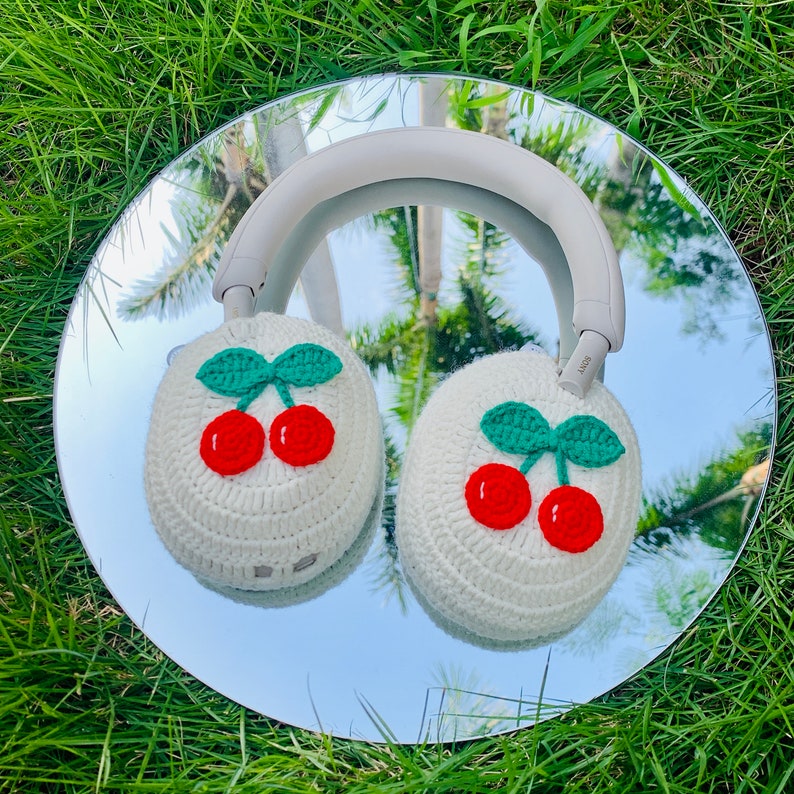 May include: White crocheted headphone covers with red cherries and green leaves. The covers are on a pair of headphones reflected in a mirror.