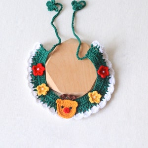 May include: A handmade crochet Christmas collar in green, white, red, and yellow. The collar features a reindeer face, flowers, and a scalloped edge. Two green ties with clover-shaped ends are attached to the collar.