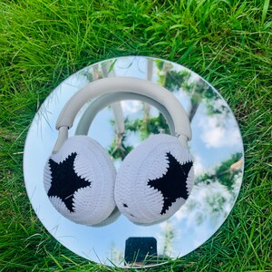May include: White knitted headphone earmuffs with black star designs. The earmuffs are on a silver reflective surface.