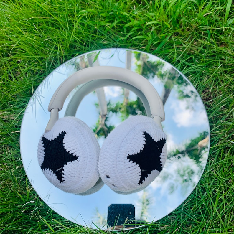 May include: White knitted headphone earmuffs with black star designs. The earmuffs are on a silver reflective surface.
