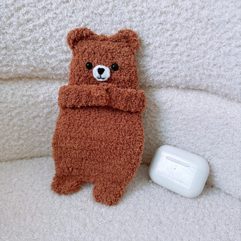 May include: A brown fuzzy bear-shaped protective case with a textured surface, featuring embroidered black eyes and a white snout with a black nose and mouth. Beside the bear case sits a smooth white rectangular wireless earbud charging case.