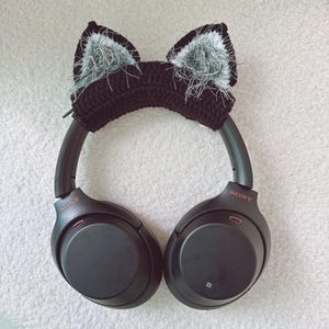 May include: Black headphones with a crocheted cat ear headband. The headband is black with white and gray fluffy accents inside the ears. The headphones have the brand name "SONY" printed on the side.