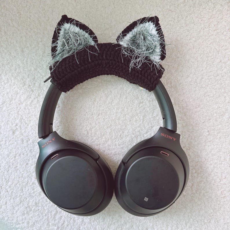 May include: Black headphones with a crocheted cat ear headband. The headband is black with white and gray fluffy accents inside the ears. The headphones have the brand name "SONY" printed on the side.