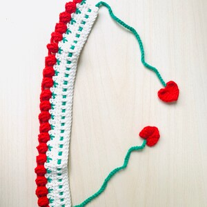 May include: A handmade crochet headband with a white base, red floral accents, and green trim. Two red heart-shaped embellishments are attached by green yarn. The headband is displayed on a light wooden surface.