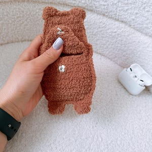 May include: A brown, fuzzy, bear-shaped pouch with a textured, fleece-like material. The pouch features a small flap secured by a silver-colored metal snap button, with a second matching snap button visible on the lower body of the bear-shaped case.
