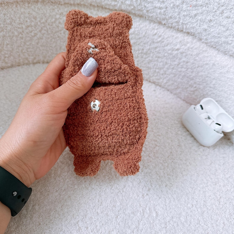 May include: A brown, fuzzy, bear-shaped pouch with a textured, fleece-like material. The pouch features a small flap secured by a silver-colored metal snap button, with a second matching snap button visible on the lower body of the bear-shaped case.