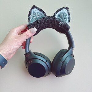 May include: Black headphones with a black crocheted cat ear headband with fluffy gray accents. The headphones are held by a hand with a blue watch. The brand name "SONY" is visible on the headphones.