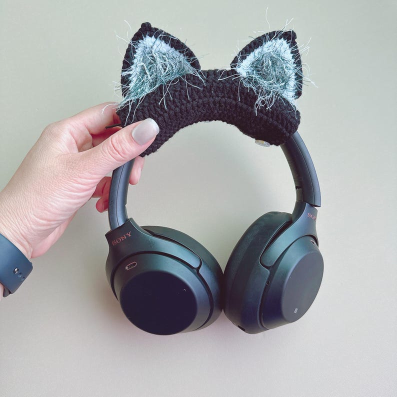 May include: Black headphones with a black crocheted cat ear headband with fluffy gray accents. The headphones are held by a hand with a blue watch. The brand name "SONY" is visible on the headphones.