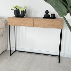 Könnte beinhalten: Ein Konsolentisch aus Holz mit zwei Schubladen und schwarzen Metallbeinen. Der Tisch ist mit einer schwarzen Vase, einer Kerze und einer Vase mit grünen Blumen dekoriert.
