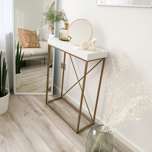 Op de afbeelding: Een witte consoletafel met een gouden metalen frame. De tafel heeft een wit blad en een gouden metalen onderstel. Er staan decoratieve items op de tafel, waaronder een vaas met gedroogde bloemen, een ronde spiegel en een klein wit beeldje. De tafel staat in een kamer met witte muren en een houten vloer.