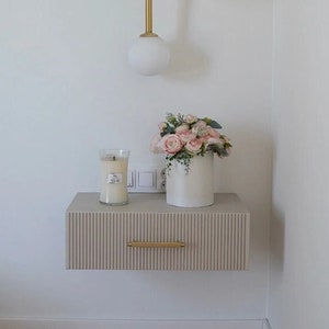 Könnte beinhalten: Eine moderne Wandleuchte mit drei weißen Kugelleuchten und goldfarbenem Finish. Darunter ein schwebender Nachttisch mit geripptem Design, goldenem Griff, einer Kerze und einer Vase mit rosa Rosen. Wohnkultur.