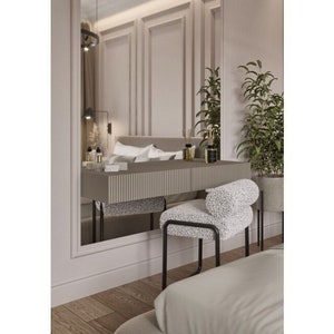 May include: A white vanity stool with a fluffy, white, textured cushion sits in front of a vanity table with a gray top and black metal legs. The vanity table is in front of a large mirror reflecting a bedroom with white walls and a white bed.