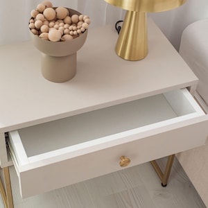 Op de afbeelding: Een wit nachtkastje met een gouden metalen frame en een enkele lade. De lade is open en de binnenkant is wit. Een gouden tafellamp met een paddenstoelvormige kap staat op het nachtkastje. Een kleine beige kom met houten ballen staat op het nachtkastje.