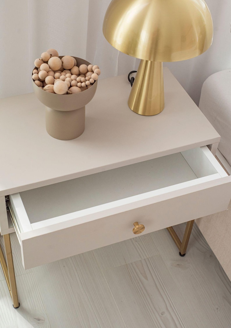 Op de afbeelding: Een wit nachtkastje met een gouden metalen frame en een enkele lade. De lade is open en de binnenkant is wit. Een gouden tafellamp met een paddenstoelvormige kap staat op het nachtkastje. Een kleine beige kom met houten ballen staat op het nachtkastje.