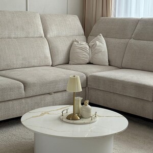 Könnte beinhalten: Ein modernes Wohnzimmer mit einem beigefarbenen Ecksofa und einem runden Couchtisch. Der Tisch hat eine weiße Platte mit goldfarbenen Adern und einen weißen Sockel. Ein Tablett mit einer Lampe, einer Kanne und einem dekorativen Glas steht auf dem Tisch.