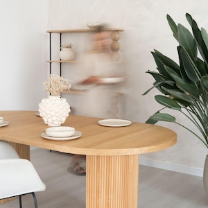 Peut inclure: Table de salle à manger ovale en bois clair avec une base cannelée, garnie d'assiettes blanches et d'un vase décoratif. Une étagère assortie et une grande plante en pot sont en arrière-plan. La table mesure environ 183 cm de long.