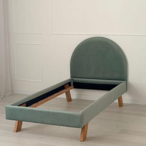 Op de afbeelding: Een saliegroen gestoffeerd bedframe met een afgeronde hoofdbord. Het frame heeft een rechthoekige vorm met houten poten. Het bedframe is ontworpen voor een eenpersoonsbed, ongeveer 190 cm lang.