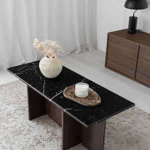 Peut inclure: Table basse rectangulaire moderne avec un plateau en marbre noir et des pieds en bois brun foncé. Une bougie blanche est posée sur un plateau décoratif. Un vase avec des fleurs séchées est sur la table.