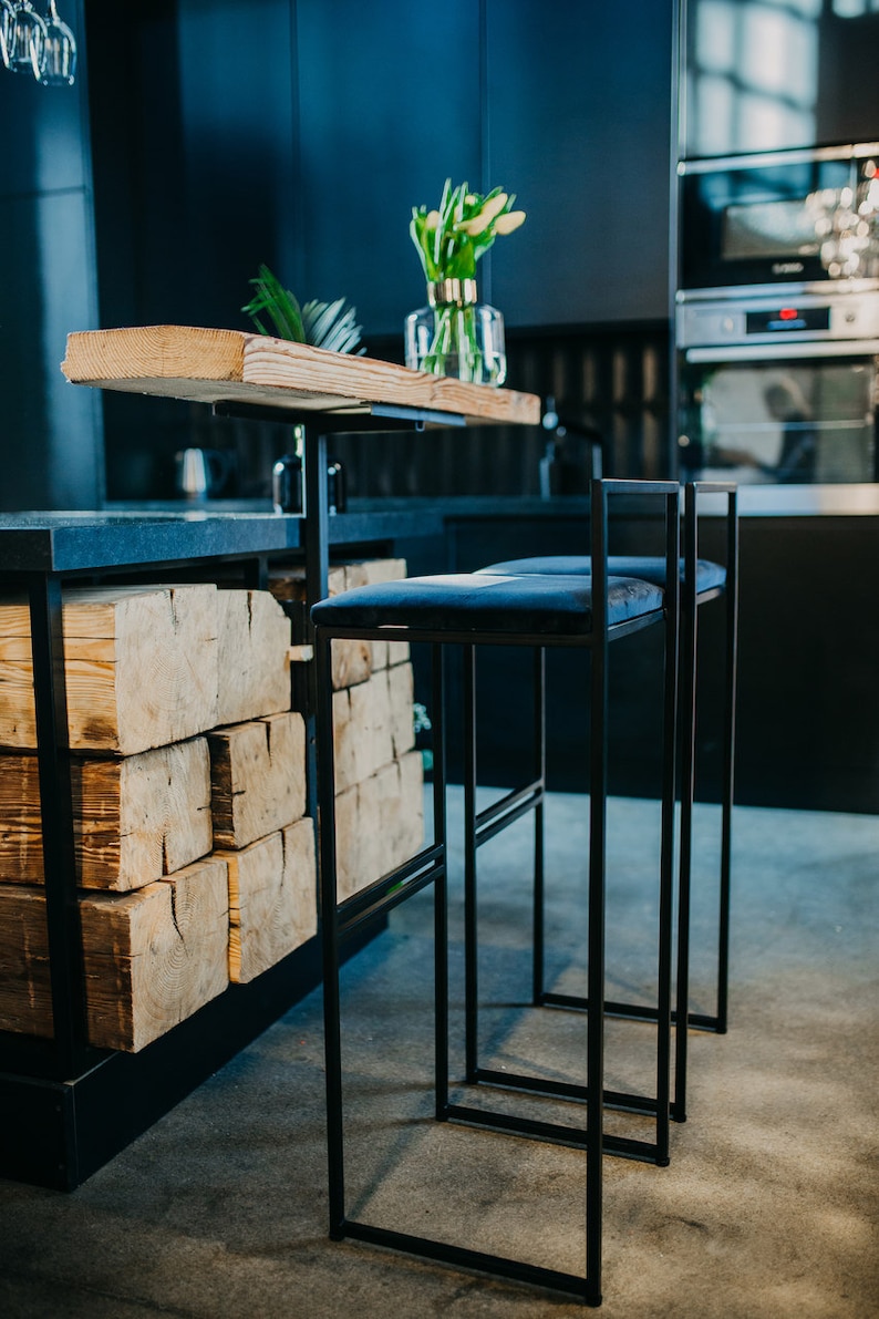 May include: Two black metal bar stools with blue velvet seats. The stools are in front of a kitchen counter with a wooden top and a black base. The counter is made of stacked wooden beams.
