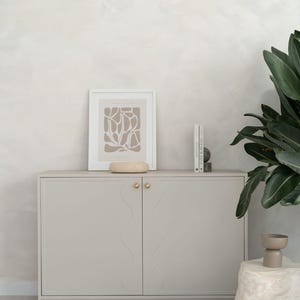 Peut inclure: Un buffet gris clair avec des boutons dorés et un motif ondulé sur les portes. Une affiche encadrée avec un motif abstrait beige, un petit bol beige et deux livres sont posés dessus. Une pierre décorative et une plante sont également visibles.