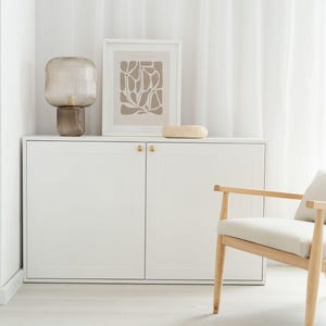 Könnte beinhalten: Moderne Innenraumgestaltung mit einem weißen Schrank, goldenen Griffen, einem gerahmten abstrakten Druck, einem dekorativen Stein und einer Glaslampe. Rechts befindet sich ein Holzstuhl mit cremefarbenem Kissen. Der Raum hat weiße Wände und einen weißen Boden.