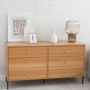 Può includere: Una cassettiera a sei cassetti con finitura in legno chiaro e gambe in metallo nero. La cassettiera ha pomelli in ottone ed è sormontata da un vaso bianco, un oggetto decorativo, una stampa incorniciata e una lampada in vetro.