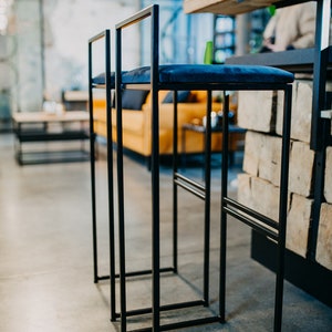 May include: Two black metal bar stools with blue velvet seats. The stools are in a modern industrial setting.