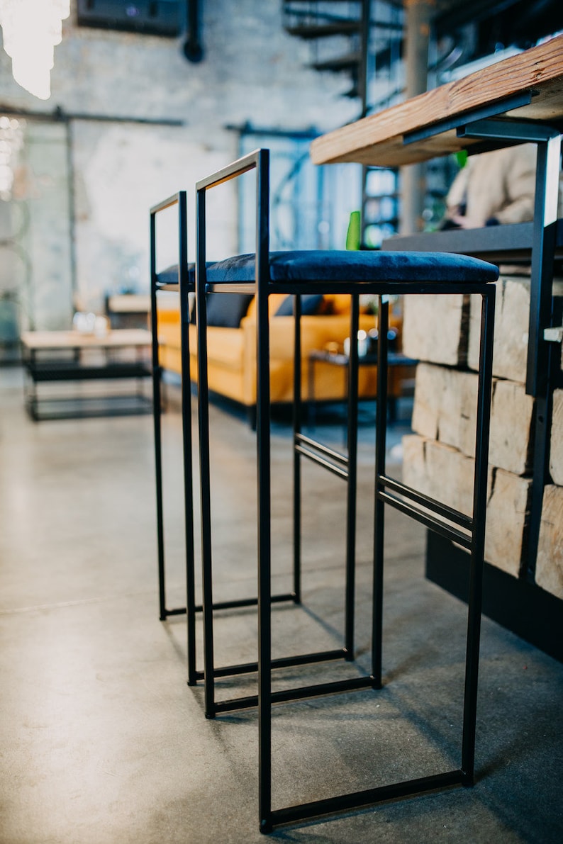 May include: Two black metal bar stools with blue velvet seats. The stools are in a modern industrial setting.