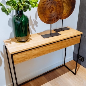 Könnte beinhalten: Ein Konsolentisch aus Holz mit einem schwarzen Metallrahmen und einer Schublade. Auf dem Tisch stehen zwei runde Wanddekorationen mit Holzmaserung. Eine grüne Glasvase mit grünen, blättrigen Pflanzen steht auf dem Tisch.