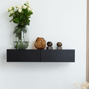 Könnte beinhalten: Ein schwarzes, geriffeltes Wandregal zeigt eine klare Glasvase mit weißen Rosen und drei dekorative Steinkugeln. Das Regal ist an einer weißen Wand befestigt und schafft eine minimalistische Ästhetik.