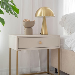 Op de afbeelding: Een witte nachtkastje met een gouden metalen frame en een lade. Op het nachtkastje staat een gouden lamp met een paddenstoelvormige kap en een decoratieve kom.