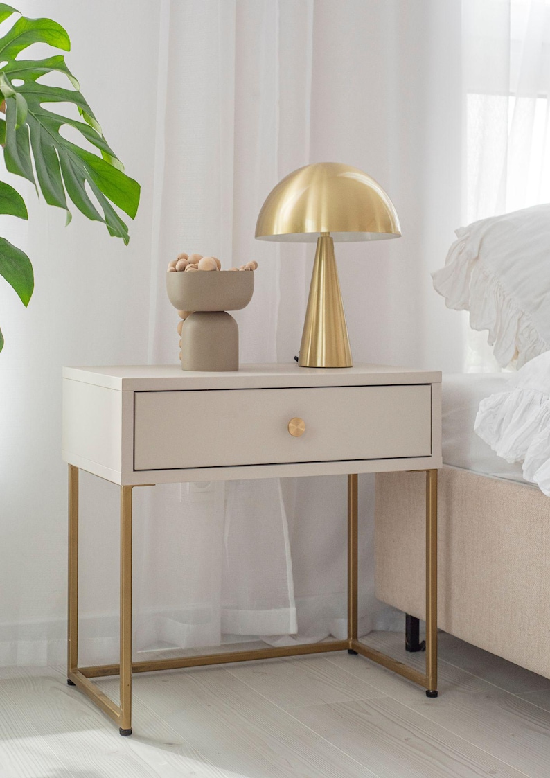 Op de afbeelding: Een witte nachtkastje met een gouden metalen frame en een lade. Op het nachtkastje staat een gouden lamp met een paddenstoelvormige kap en een decoratieve kom.