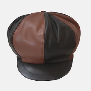 Puede incluir: Gorra de estilo newsboy bicolor en cuero marrón y negro. La gorra presenta una corona redondeada y una visera corta y curvada. El diseño de bloques de color añade un toque moderno al estilo clásico. Este sombrero es un accesorio elegante.