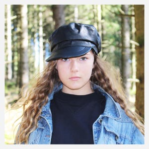 Può includere: Una persona giovane con i capelli lunghi e ricci che indossa un cappello da newsboy in pelle nera e una giacca di jeans blu.