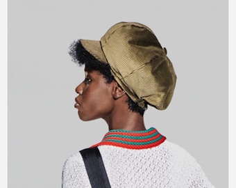 May include: A person wearing a white knitted sweater with red and green stripes around the collar, a brown corduroy newsboy cap, and a black strap across their shoulder.
