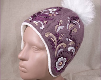 May include: A purple and white embroidered winter hat with a white faux fur pom pom. The hat has a white fleece lining.