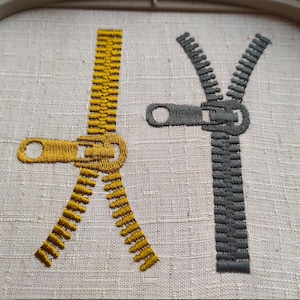 May include: Two embroidered zippers, one gold and one gray, on a white fabric background.