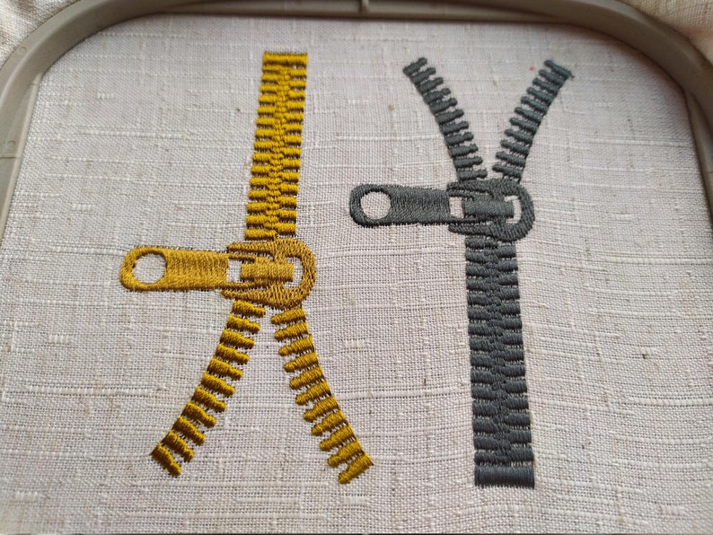 May include: Two embroidered zippers, one gold and one gray, on a white fabric background.