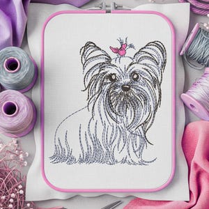 K&ouml;nnte beinhalten: Gesticktes Design eines Yorkshire Terriers in einem rosa Stickrahmen. Der Hund hat langes, flie&szlig;endes Fell in Grau- und Schwarzt&ouml;nen, mit einem rosa Schmetterling auf dem Kopf. Der Hintergrund umfasst Garnrollen und Stoff.