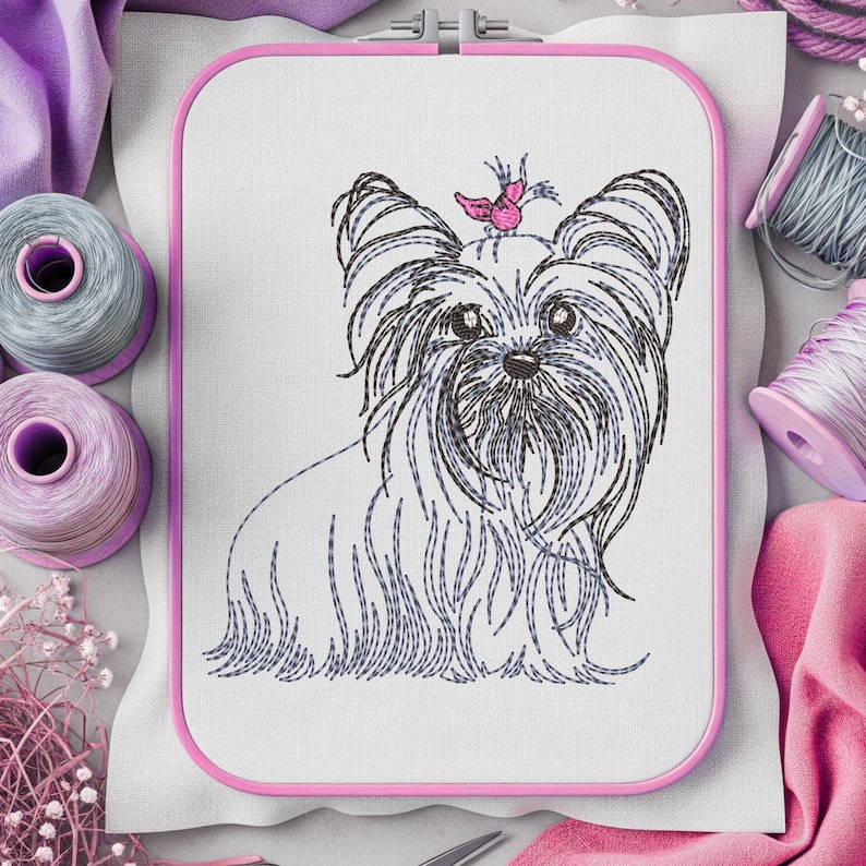 K&ouml;nnte beinhalten: Gesticktes Design eines Yorkshire Terriers in einem rosa Stickrahmen. Der Hund hat langes, flie&szlig;endes Fell in Grau- und Schwarzt&ouml;nen, mit einem rosa Schmetterling auf dem Kopf. Der Hintergrund umfasst Garnrollen und Stoff.