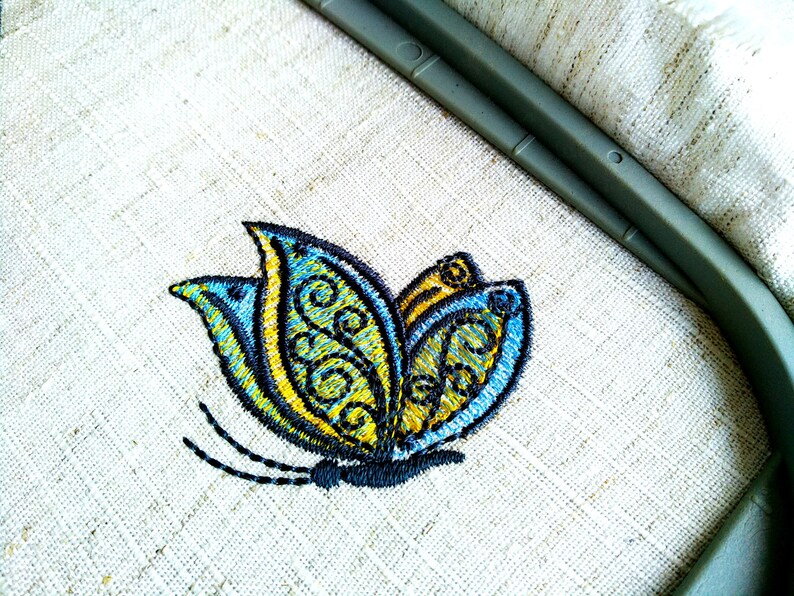 May include: A blue and yellow embroidered butterfly design on a white linen fabric.