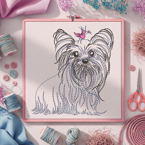 K&ouml;nnte beinhalten: Gerahmte Stickerei eines Yorkshire Terriers mit schwarzen und blauen Fadendetails. Der Hund hat eine rosa Schleife auf dem Kopf. Die Stickerei befindet sich in einem rosa Rahmen, mit Garnrollen, Kn&ouml;pfen und einer Schere im Hintergrund.
