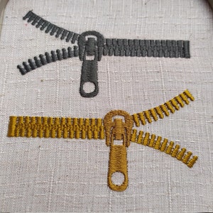 May include: Two embroidered zippers, one in black and one in gold, are shown on a white fabric background. The zippers are open and the pull tabs are visible.
