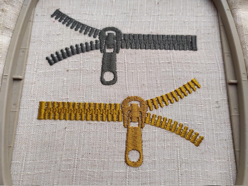 May include: Two embroidered zippers, one in black and one in gold, are shown on a white fabric background. The zippers are open and the pull tabs are visible.