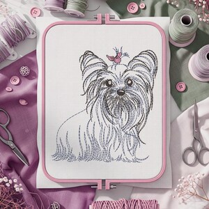 K&ouml;nnte beinhalten: Ein rosa Stickrahmen zeigt ein detailliertes Stickbild eines Yorkshire Terrier-Hundes. Der Hund ist mit schwarzem und blauem Garn gestickt, mit einer rosa Schleife. N&auml;hzubeh&ouml;r, einschlie&szlig;lich Garnrollen, Scheren und Kn&ouml;pfen, umgibt den Rahmen.