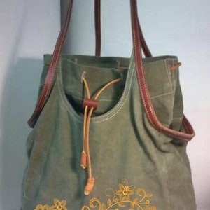 K&ouml;nnte beinhalten: Eine gr&uuml;ne Canvas-Tragetasche mit braunen Lederriemen und einem Kordelzug. Die Tasche ist mit einem goldenen, gestickten Blumendesign verziert.