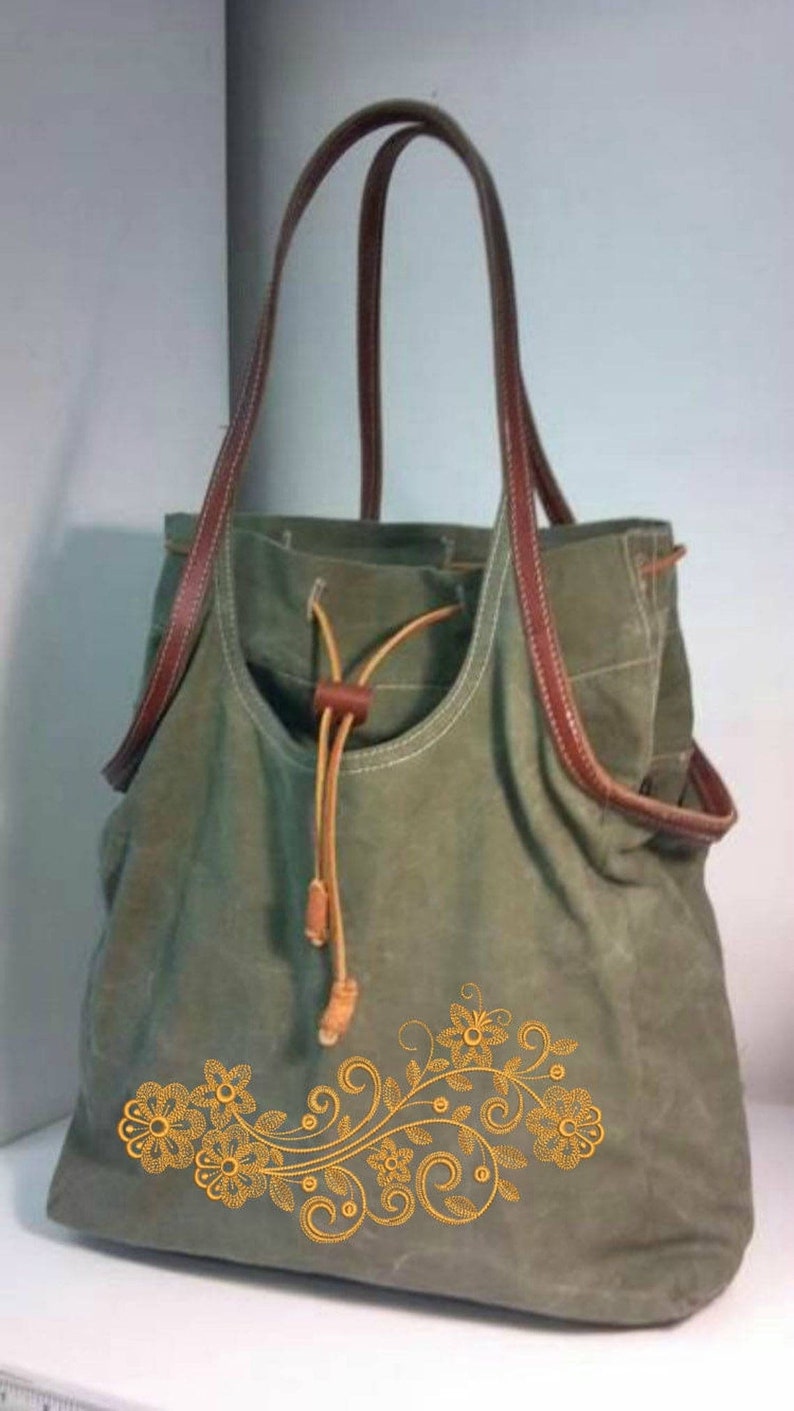K&ouml;nnte beinhalten: Eine gr&uuml;ne Canvas-Tragetasche mit braunen Lederriemen und einem Kordelzug. Die Tasche ist mit einem goldenen, gestickten Blumendesign verziert.