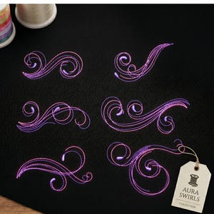 Puede incluir: Seis diseños de bordado distintos con patrones de remolinos en tonos morados y rosas cosidos sobre tela negra. Se adjunta una etiqueta de papel beige que muestra un icono de bobina y el texto AURA SWIRLS COLLECTION en letras negras.