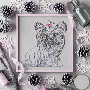K&ouml;nnte beinhalten: Gerahmte Stickerei eines Yorkshire Terriers mit einem rosa Schmetterling auf dem Kopf. Das Kunstwerk ist von dekorativen Elementen umgeben, darunter Tannenzapfen, Spitzenschneeflocken und eine Glasflasche, alles in Grau-, Wei&szlig;- und Rosat&ouml;nen.